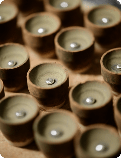 Primo piano di diversi piccoli vasi rotondi di argilla marrone disposti in fila, ognuno dei quali contiene una singola perlina o biglia d'argento lucida al centro.
