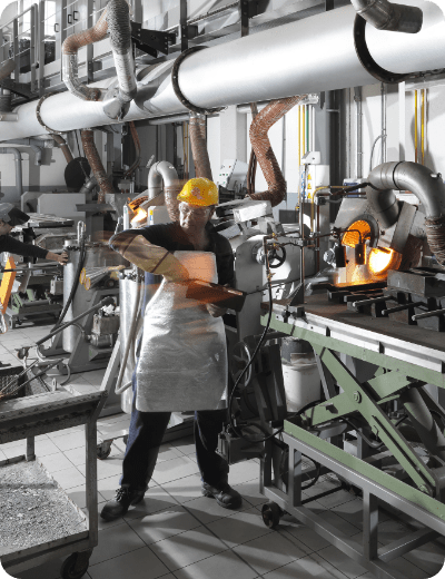 Un operaio che indossa un elmetto giallo, occhiali di sicurezza e un grembiule aziona i macchinari in un'officina industriale piena di tubi, strumenti e attrezzature.