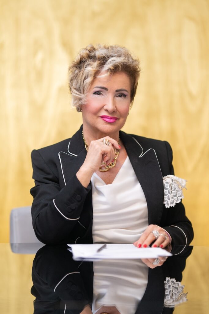 Ivana Ciabatti, fundadora y presidenta de Italpreziosi, está sentada ante un escritorio y lleva una americana negra con ribetes blancos y una blusa blanca. Sostiene un bolígrafo y un papel, con un fondo amarillo neutro detrás de ella.