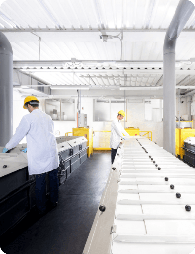 Due operai con camici bianchi e caschi gialli azionano macchinari in un moderno e luminoso impianto industriale con grandi tubi e nastri trasportatori bianchi.