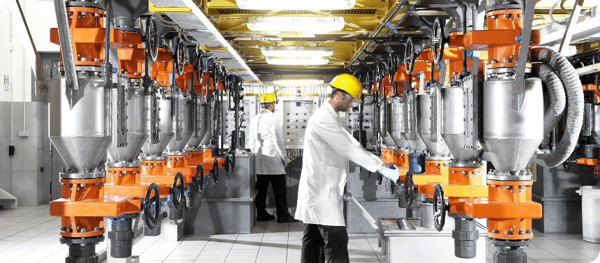 Due operai in camice da laboratorio e elmetto giallo manovrano attrezzature industriali con macchinari arancioni e argentati in un ambiente di fabbrica moderno e luminoso.