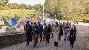 Un gruppo di persone cammina all'aperto lungo un sentiero di ghiaia, passando davanti a diverse bandiere internazionali sui pennoni. Sullo sfondo sono visibili alberi e un'area erbosa. La maggior parte delle persone indossa un cappotto.