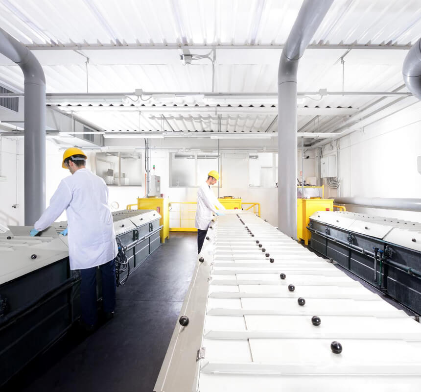 Dos trabajadores con batas blancas de laboratorio y cascos amarillos están de pie en una luminosa instalación industrial, manejando grandes máquinas rectangulares bajo un techo alto con vigas y tuberías a la vista.