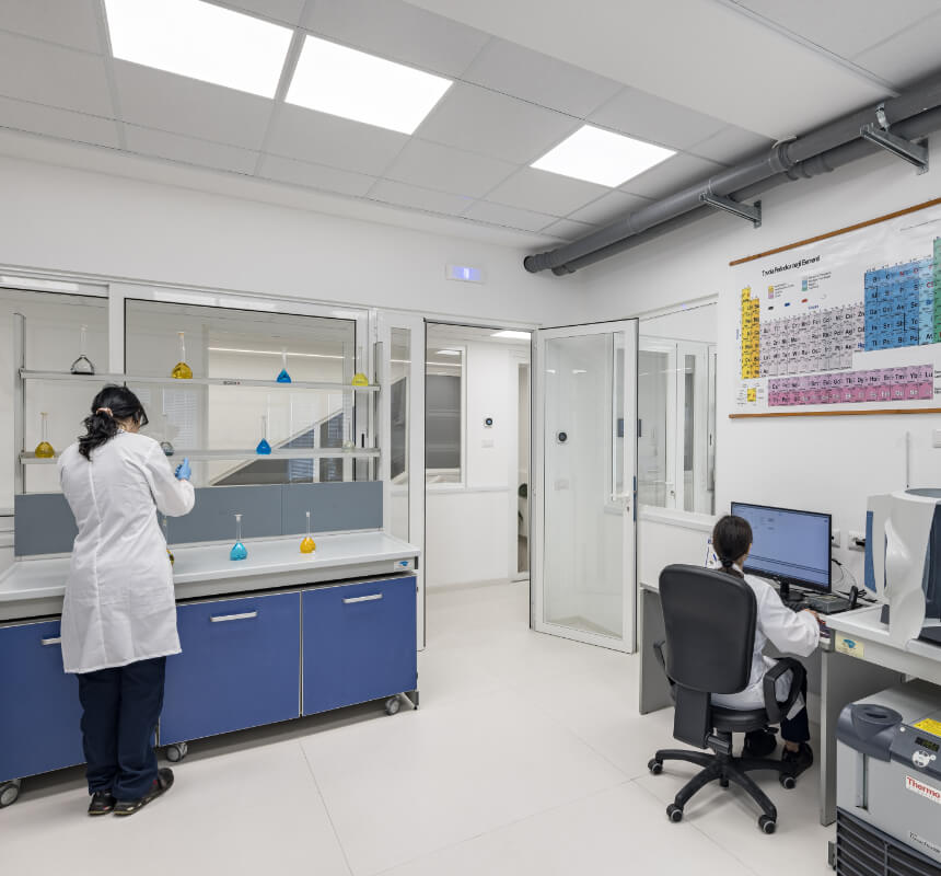 Dos personas con batas de laboratorio trabajan en un luminoso laboratorio; una está de pie junto a un mostrador manipulando equipos, mientras que la otra está sentada en un escritorio utilizando un ordenador. En la pared hay una tabla periódica.