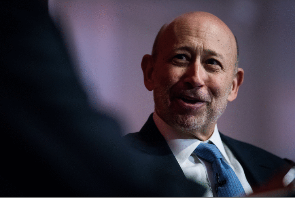 Un uomo calvo con la barba grigia, che indossa un abito e una cravatta azzurra, sorride mentre parla con qualcuno fuori campo in un ambiente poco illuminato.