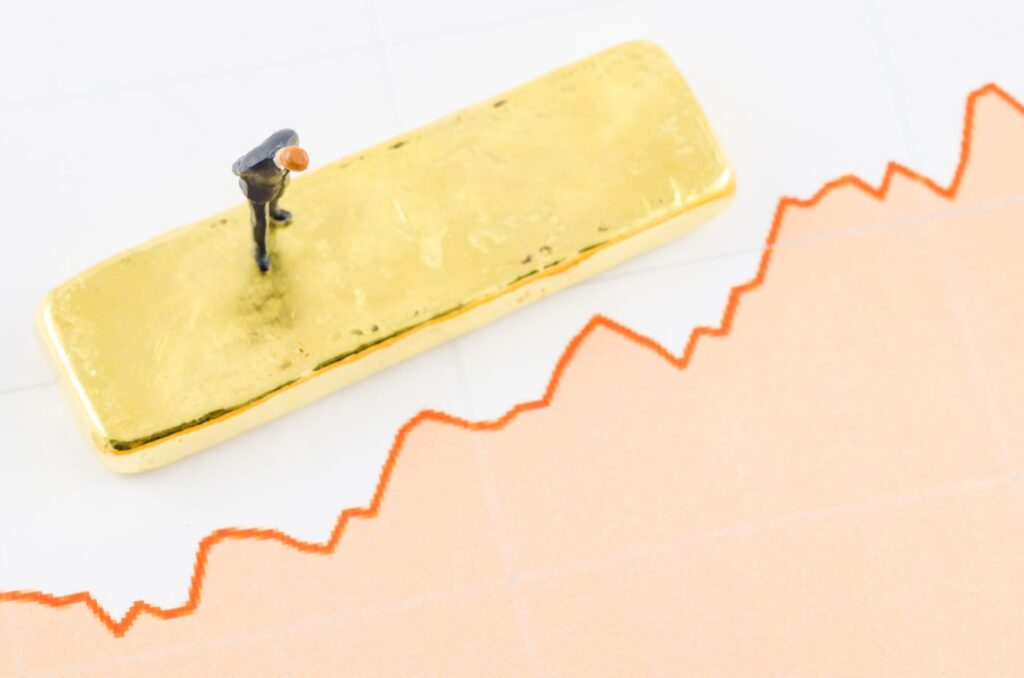 La miniatura di una persona si trova su un lingotto d'oro posto sopra un grafico finanziario con una linea di tendenza rossa ascendente, che simboleggia la crescita degli investimenti in oro.