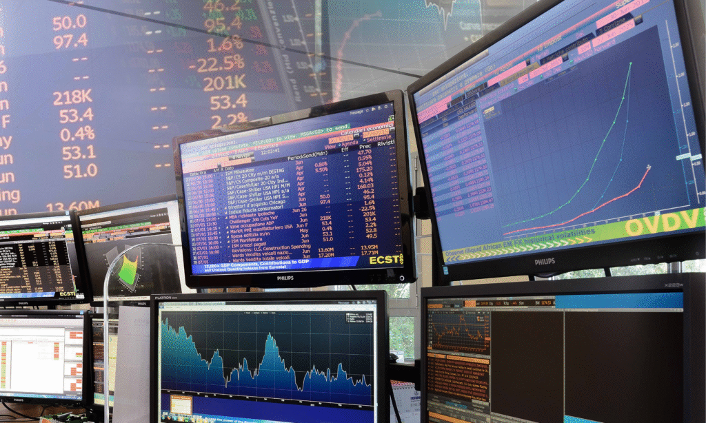 Più monitor di computer visualizzano dati finanziari, grafici e diagrammi in un ambiente di trading, con cifre colorate e informazioni sulle azioni visibili sugli schermi.