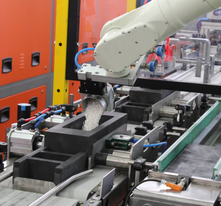 Un brazo robótico vierte pequeños gránulos de plástico blanco en un molde negro rectangular en una cadena de montaje industrial, con equipos naranjas al fondo.