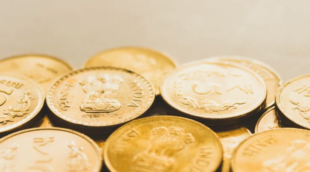 Una vista ravvicinata di diverse monete indiane, per lo più in tonalità oro, sovrapposte l'una all'altra con iscrizioni e simboli visibili, tra cui il leone capitale di Ashoka. Lo sfondo è leggermente sfocato.