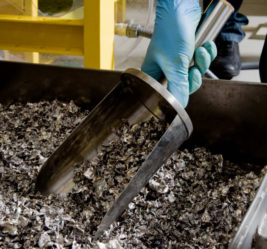 Una mano enguantada sostiene una gran pala metálica llena de material gris y escamoso, posiblemente cenizas industriales o virutas de metal, sobre un cubo. Al fondo se ven máquinas y tuberías amarillas.