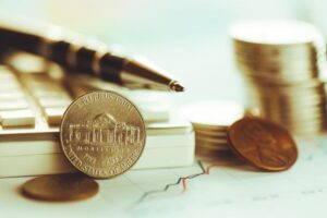 Primo piano di un nichelino statunitense in piedi su una tastiera, con una penna, monete impilate e un penny sullo sfondo, il tutto su un grafico finanziario con una linea ascendente.