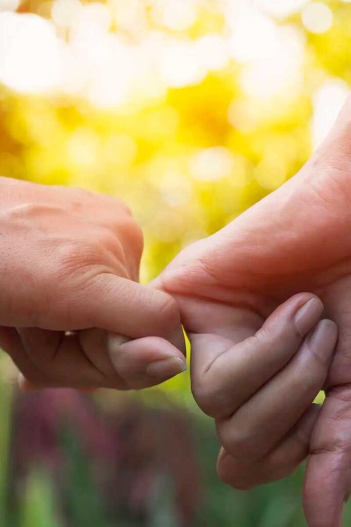 Dos manos sujetándose suavemente por los dedos meñiques, simbolizando conexión o promesa, con un fondo borroso de luz solar dorada.
