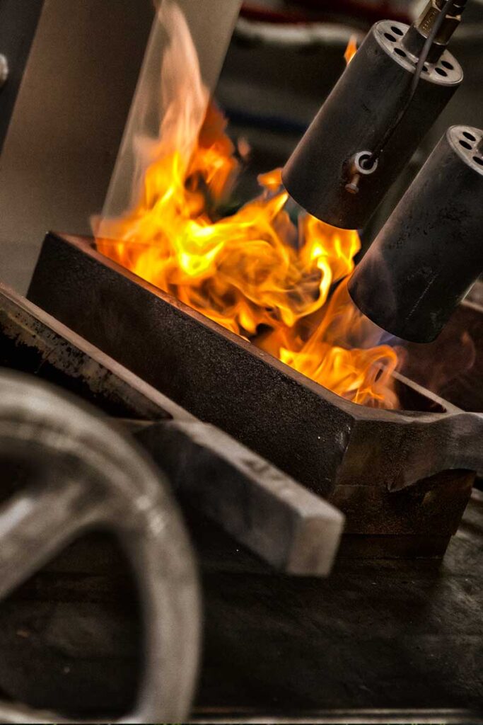 De un recipiente metálico salen llamas mientras dos sopletes cilíndricos calientan la superficie en un entorno industrial. En primer plano se ve una rueda metálica difuminada.