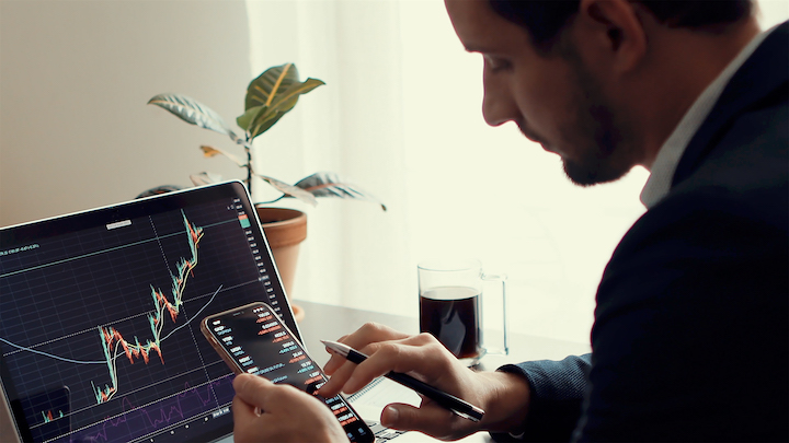 Un uomo in giacca e cravatta analizza i grafici del mercato azionario su un computer portatile e uno smartphone alla scrivania, con una tazza di caffè, una penna e una pianta in vaso nelle vicinanze. Sullo schermo sono visualizzati grafici finanziari con tendenza al rialzo.