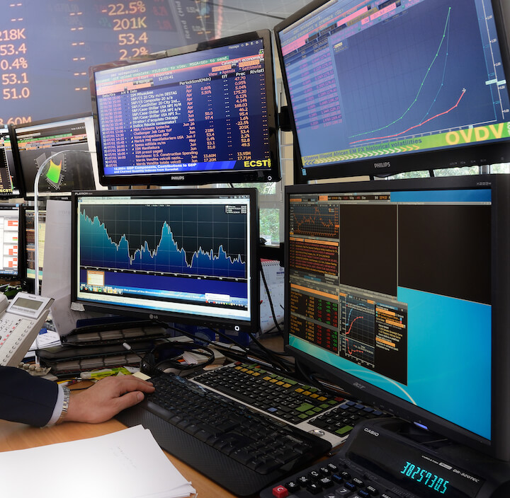 Una persona lavora a una scrivania con diversi monitor di computer che visualizzano grafici azionari, dati finanziari e grafici in un ambiente di trading affollato. Una mano è sulla tastiera e i fogli sono impilati nelle vicinanze.