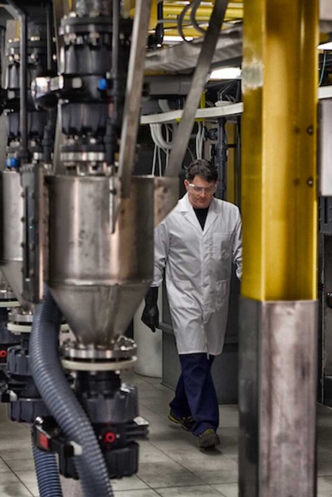 Una persona con bata blanca de laboratorio y gafas de seguridad camina por una instalación industrial con grandes máquinas y tuberías de acero inoxidable.