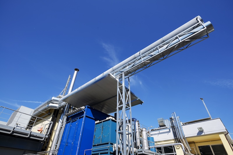 Instalación industrial con grandes tuberías metálicas, conductos y maquinaria sobre un cielo azul despejado. En la estructura destaca una tubería plateada en ángulo sostenida por una estructura metálica sobre otros equipos.