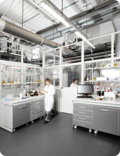 Dos personas con bata blanca trabajan en un moderno laboratorio con armarios de acero inoxidable, equipos de laboratorio y estanterías con botellas y cristalería. El espacio tiene grandes ventanales y tuberías a la vista.