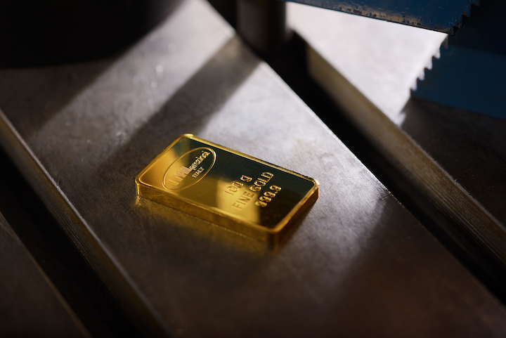 Un singolo lingotto d'oro poggia su una superficie metallica sotto una luce intensa, mostrando la sua finitura lucida e riflettente e i dettagli incisi, tra cui le indicazioni di peso e purezza.