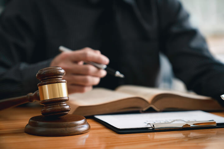 Una persona in abiti scuri siede a una scrivania con un martelletto, un libro aperto, una cartellina e una penna, suggerendo un ambiente legale o giudiziario.