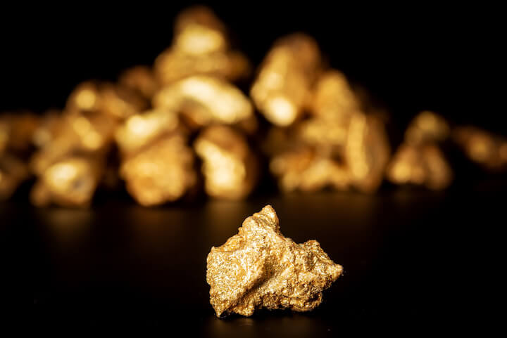 Una pepita d'oro lucente è messa a fuoco in primo piano, con un mucchio sfocato di altre pepite d'oro sullo sfondo su una superficie nera.