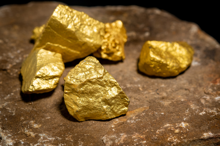 Pezzi di pepite d'oro lucenti appoggiati su una superficie di pietra grezza e marrone, con uno sfondo scuro.