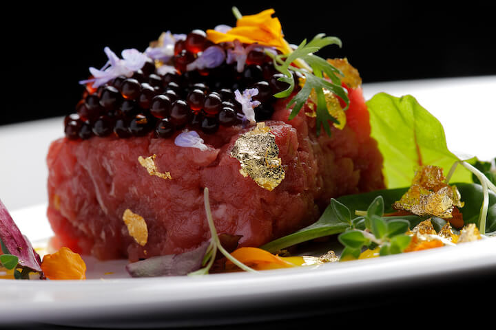 Primo piano di una steak tartare gourmet condita con caviale nero, foglie d'oro commestibili e delicati fiori viola, servita con microgreens e petali commestibili colorati su un piatto bianco su sfondo nero.