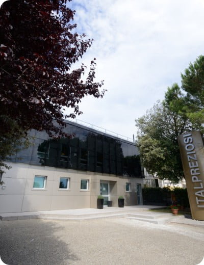 Un edificio moderno con grandes ventanas tintadas de oscuro y paredes de color claro, rodeado de árboles. Un cartel alto en primer plano indica ITALPREZIOSI. El cielo está parcialmente nublado.