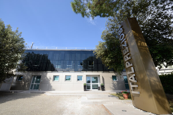 Moderno edificio con facciata in vetro, alberi su entrambi i lati e una grande insegna verticale con la scritta ITALPREZIOSI in caratteri dorati vicino all'ingresso, sotto un cielo azzurro e limpido.
