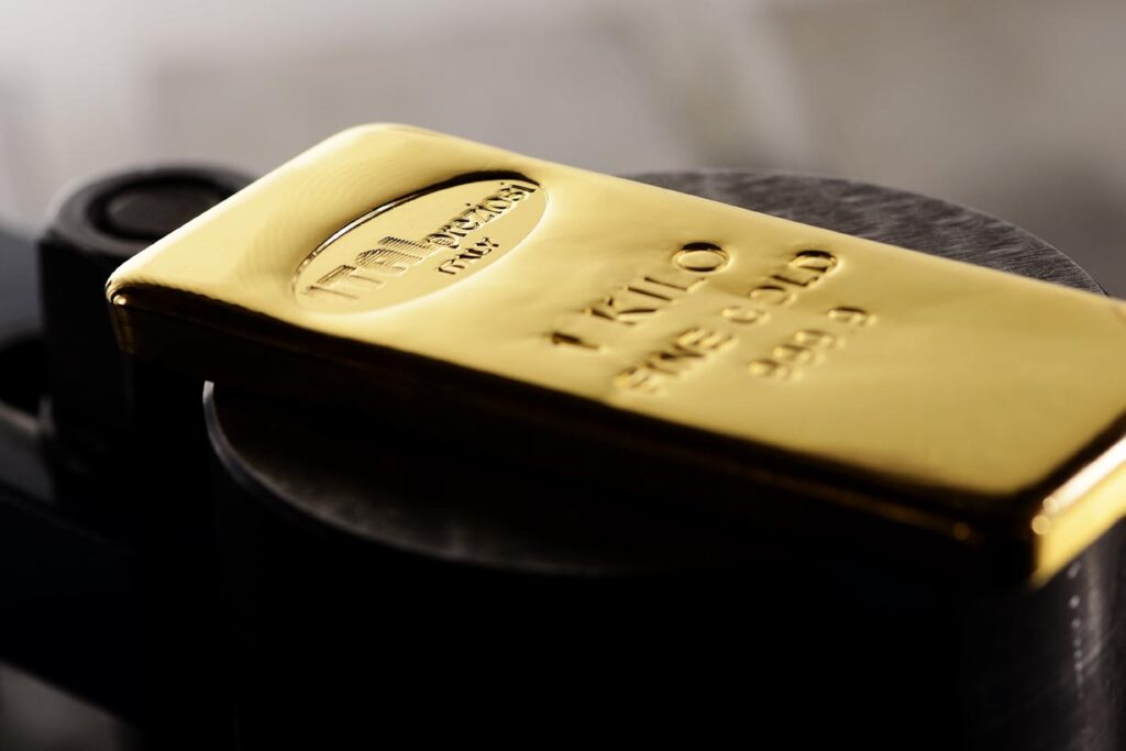 Primo piano di un lingotto d'oro lucido con la dicitura 1 KILO FINE GOLD 999.9, appoggiato su una superficie metallica scura. Il lingotto d'oro riflette la luce, evidenziando la sua consistenza liscia e il suo colore ricco.