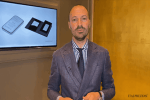 Un uomo in abito blu si trova davanti a una parete gialla e a uno schermo che mostra immagini di barrette e confezioni d'argento, con il logo italpreziosi visibile in un angolo.