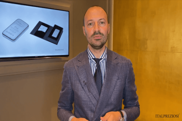 Un uomo in abito blu si trova davanti a una parete gialla e a uno schermo che mostra immagini di barrette e confezioni d'argento, con il logo italpreziosi visibile in un angolo.