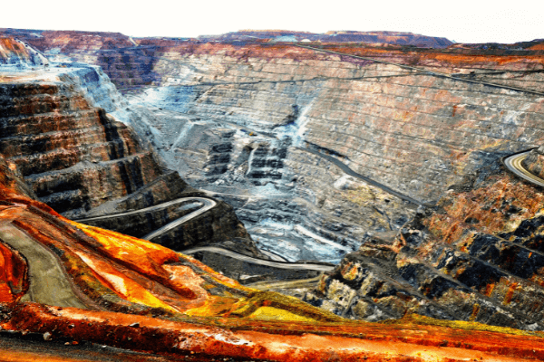 Una grande miniera a cielo aperto con ripide pareti di roccia terrazzate e strade tortuose che scendono nel pozzo. La terra esposta mostra strati di tonalità rosse, arancioni e grigie sotto un cielo luminoso.