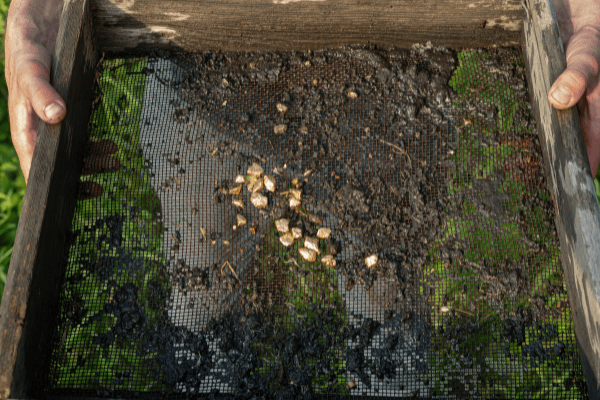 Una persona tiene in mano un paravento con struttura in legno e rocce, terra e piccoli manufatti o punte di freccia esposti sulle maglie, probabilmente provenienti da uno scavo archeologico o da un'attività di setaccio all'aperto.