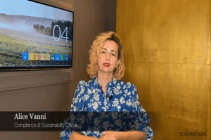 Una donna con capelli biondi corti e ricci e rossetto rosso, che indossa una camicetta blu a fiori, siede in casa. Alle sue spalle uno schermo a parete e un muro giallo. Il testo recita Alice Vanni, Compliance Sustainability Officer, Italpreziosi.