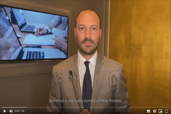 Un uomo in giacca e cravatta si trova al chiuso, di fronte alla telecamera, con un microfono agganciato alla giacca. Uno schermo televisivo alle sue spalle mostra persone che lavorano su computer portatili e documenti. I sottotitoli in italiano sono visibili in basso.