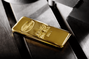 Un lingotto d'oro lucido con la scritta "1 KILO FINE GOLD 999.9" poggia su una superficie metallica, parzialmente illuminata dalla luce, con incisioni e riflessi visibili.