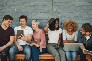 Sei giovani adulti siedono su una panchina contro un muro di mattoni, sorridendo e ridendo mentre guardano tablet e computer portatili, suggerendo un'atmosfera informale, amichevole e collaborativa.
