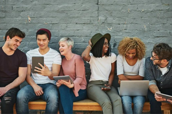 Sei giovani adulti siedono su una panchina contro un muro di mattoni, sorridendo e ridendo mentre guardano tablet e computer portatili, suggerendo un'atmosfera informale, amichevole e collaborativa.