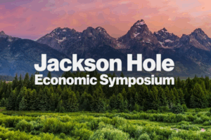 Text Jackson Hole Economic Symposium over a scenic landscape with dense green trees in the foreground and rugged mountain peaks beneath a colorful sky in the background.
