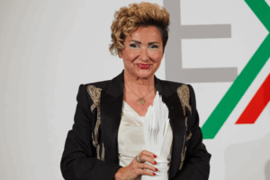 Ivana Ciabatti, fondatrice e presidente di Italpreziosi, sorride tenendo in mano un trofeo bianco davanti a uno sfondo bianco con forme geometriche verdi e rosse.