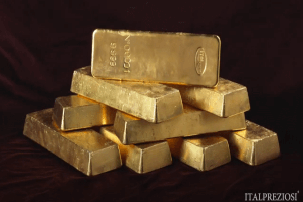 Una pila di lingotti d'oro lucidi è disposta su una superficie scura, con un lingotto posto in cima. I lingotti d'oro presentano iscrizioni e riflettono la luce, evidenziando la loro consistenza metallica. La parola "ITALPREZIOSI" appare nell'angolo in basso a destra.