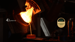 Il metallo fuso viene versato da una fornace in uno stampo circolare in un ambiente industriale scarsamente illuminato, con il logo Italpreziosi e il badge verde del premio di sostenibilità visibili sui lati sinistro e destro dell'immagine.