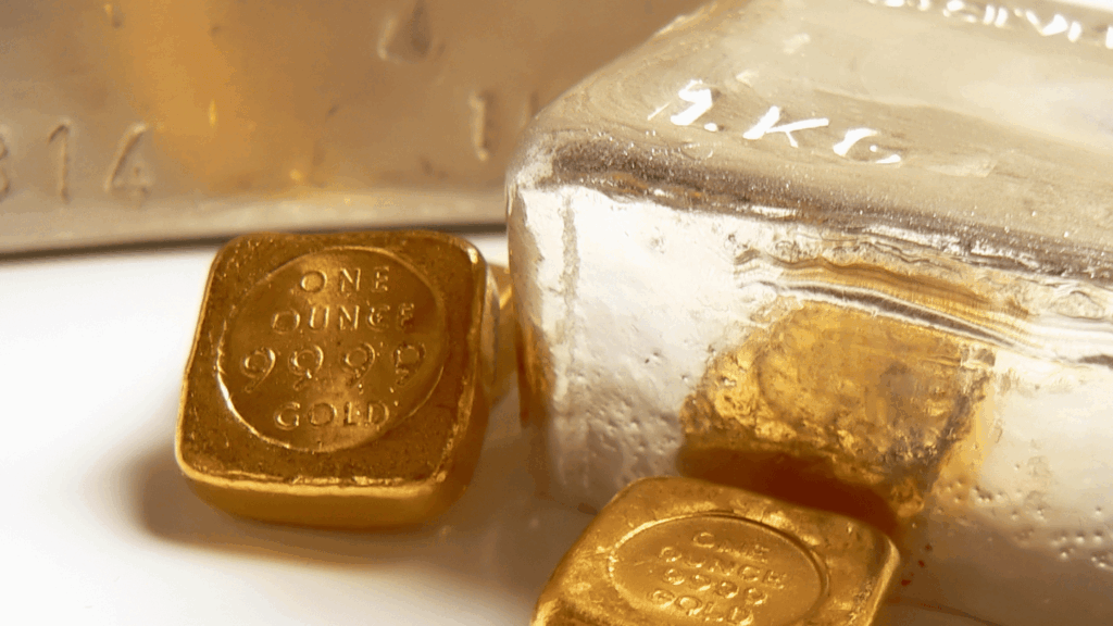 Primo piano di lingotti d'oro con l'etichetta "ONE OUNCE 999.9 GOLD" accanto a un lingotto d'argento più grande con l'etichetta "1 KG". Le superfici sono lucide e riflettono la luce.
