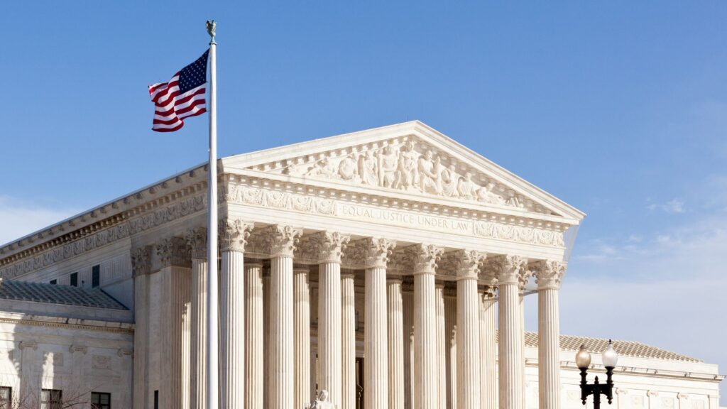 L'edificio della Corte Suprema degli Stati Uniti con alte colonne, intagli ornamentali e una bandiera americana che sventola in primo piano in un cielo azzurro e limpido.
