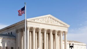 L'edificio della Corte Suprema degli Stati Uniti con alte colonne, intagli ornamentali e una bandiera americana che sventola in primo piano in un cielo azzurro e limpido.