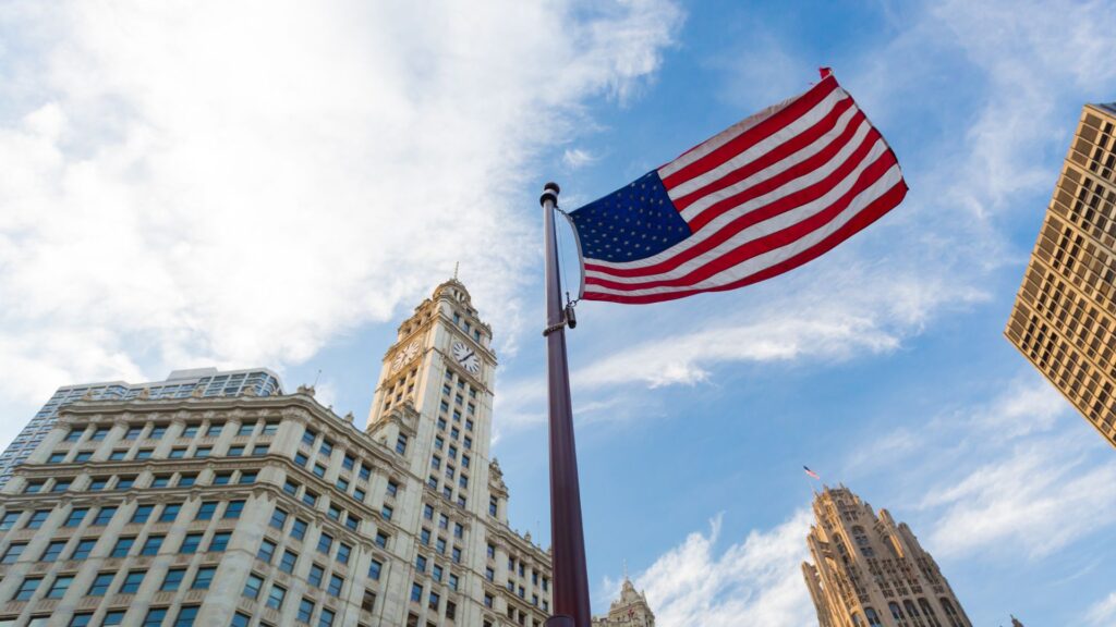 Una bandiera americana sventola su un'asta in un cielo blu con nuvole, circondata da alti edifici storici in un paesaggio urbano.