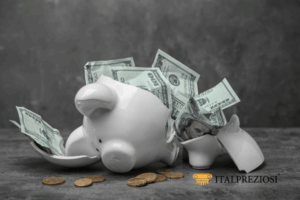 Un salvadanaio bianco rotto con banconote e monete da un dollaro USA che fuoriescono, posto su una superficie grigia con sfondo scuro. Il logo Italpreziosi si trova nell'angolo in basso a destra.