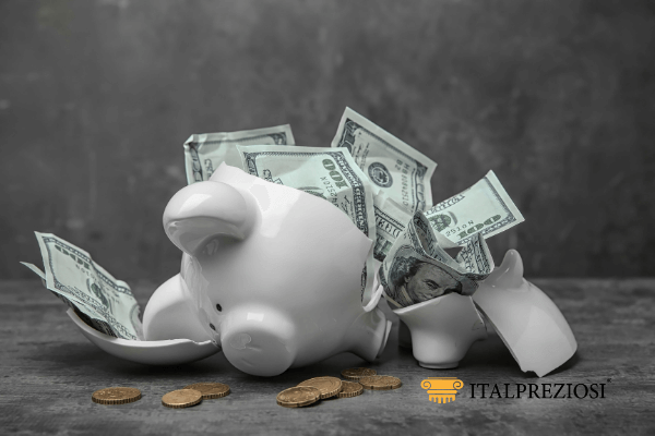 Un salvadanaio bianco rotto con banconote e monete da un dollaro USA che fuoriescono, posto su una superficie grigia con sfondo scuro. Il logo Italpreziosi si trova nell'angolo in basso a destra.
