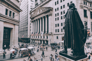 Scena di strada affollata nel Financial District di New York, con una grande statua in primo piano e l'edificio della Borsa di New York con bandiere americane sullo sfondo, circondato da persone e venditori.
